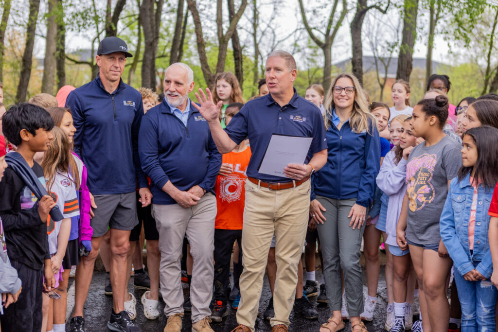 Man holding a document speaks to a group of schoolchildren and adults gathered outdoors among trees.