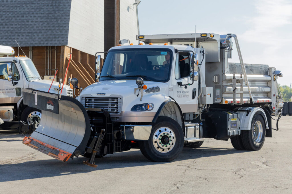 White New Albany Public Service snowplow truck parked, large curved metal plow with orange edge and a rear dump bed
