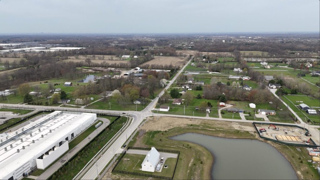 Aerial view of rural area with intersection, large white warehouse, fenced house by a pond and scattered homes