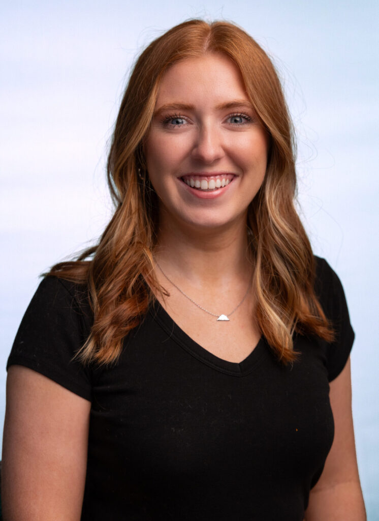 Smiling woman with wavy red hair and blue eyes wearing a black V-neck and small pendant against a light background.