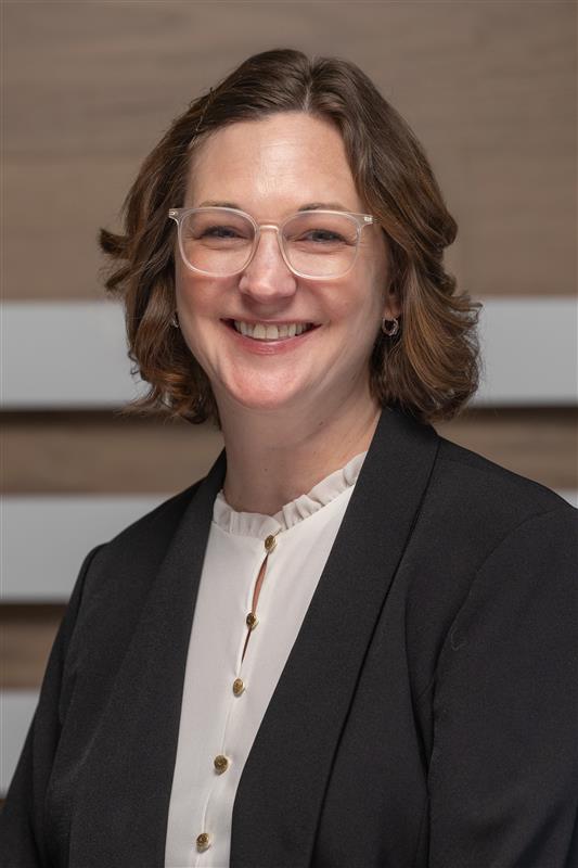 Smiling woman with short wavy brown hair and clear glasses wearing a black blazer and white blouse against wood background