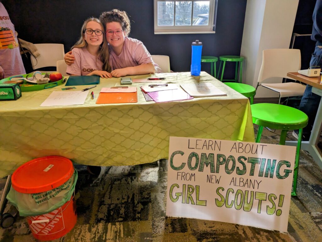 Two smiling girls at a composting booth with clipboards and a sign that reads Learn about composting from local Girl Scouts