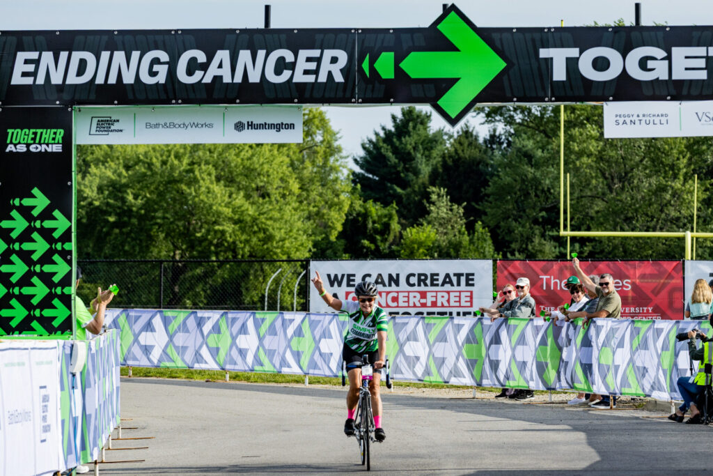 Cyclist in green jersey raises an arm crossing a finish line under a large ENDING CANCER banner while spectators cheer