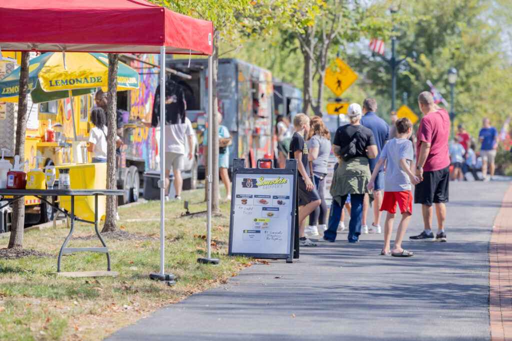 People lined up along a tree-lined sidewalk at food trucks and a lemonade stand with yellow umbrellas and a red canopy.