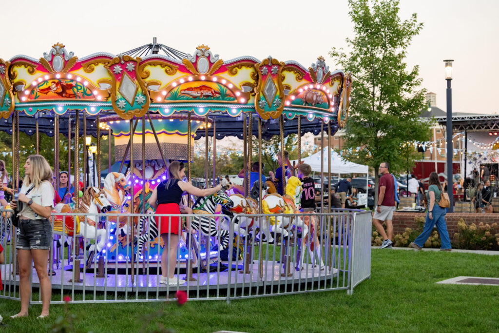 Colorful lit carousel with painted horses and children riding, surrounded by adults at an outdoor evening fair.