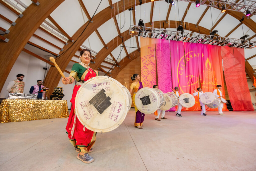 Woman in green-and-red sari playing a large dhol drum leading a line of drummers on a colorful Celebrate Diwali stage.