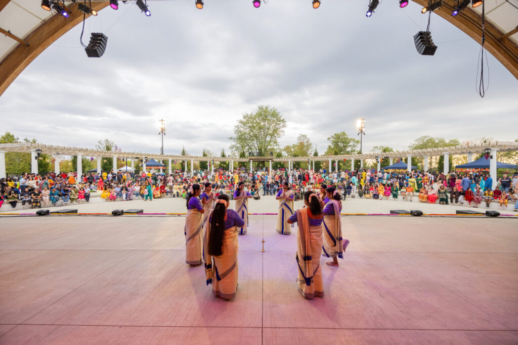 Group of women in cream-and-blue sarees performing a circular dance on an outdoor stage with a large seated audience.
