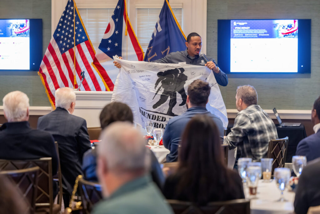 Presenter holding a signed Wounded Warrior Project flag with soldier silhouette as seated audience watches in a meeting room.