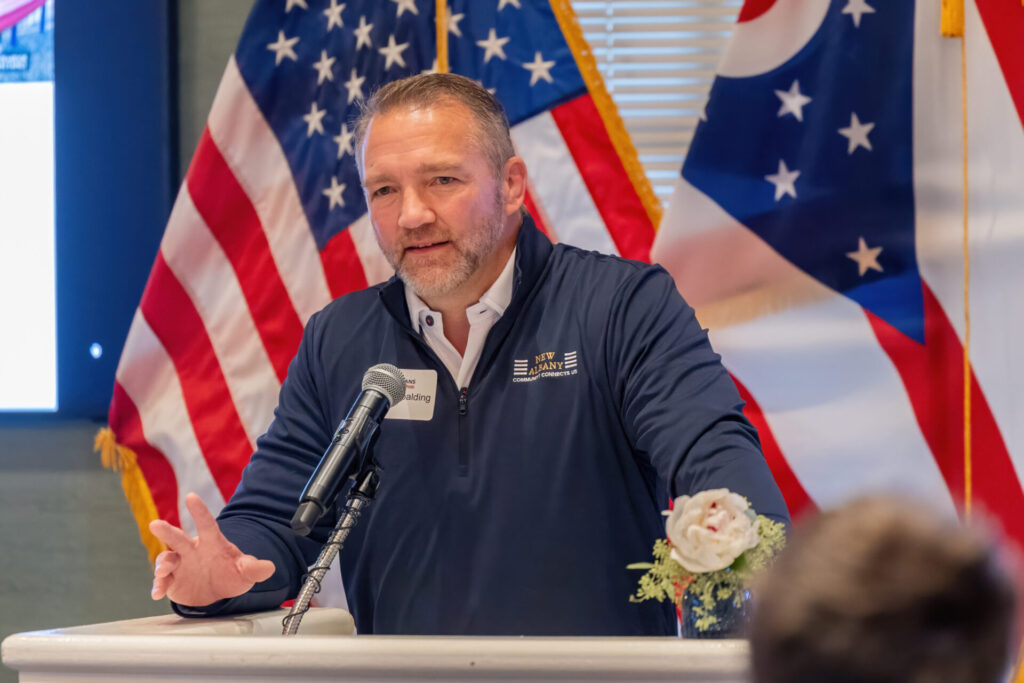 Man speaking at a podium with microphone, wearing a navy New Albany pullover, U.S. flags behind him.