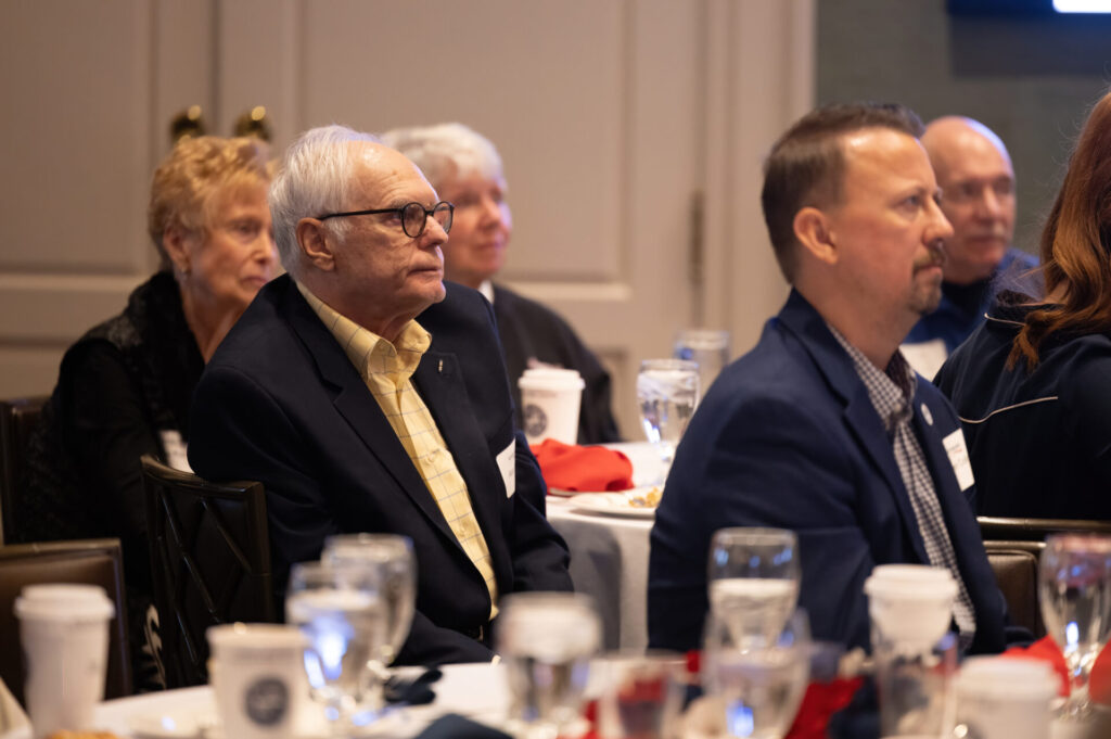 Elderly man with round glasses in a dark jacket and yellow checked shirt seated at a banquet table listening attentively.