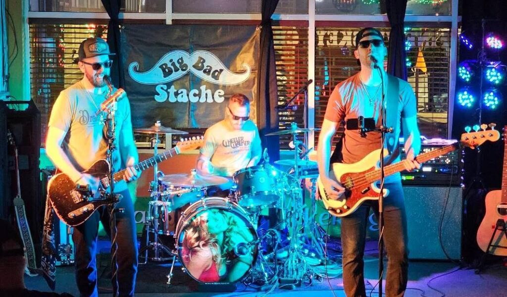 Three-piece band playing under colorful stage lights with a Big Bad Stache banner, two guitarists and a drummer.