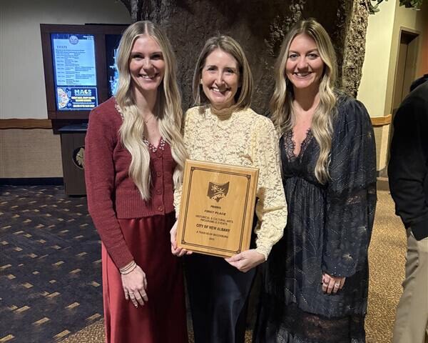 Three smiling women indoors by a tree trunk; the center woman in cream lace holds a wooden first-place plaque, flanked by women in red and black.