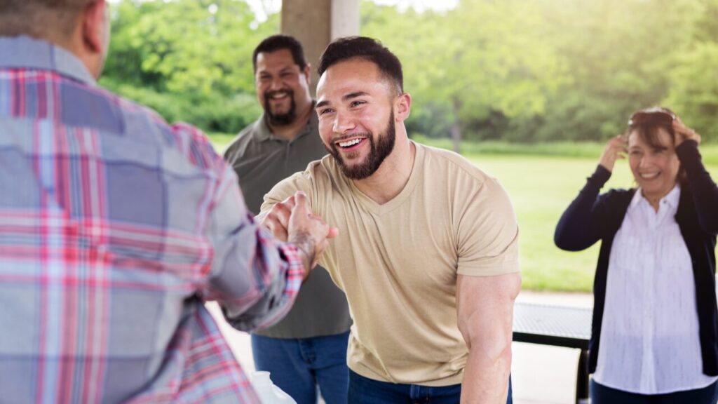 Young bearded man in beige shirt shaking hands with someone in plaid under a park shelter as friends smile behind him.