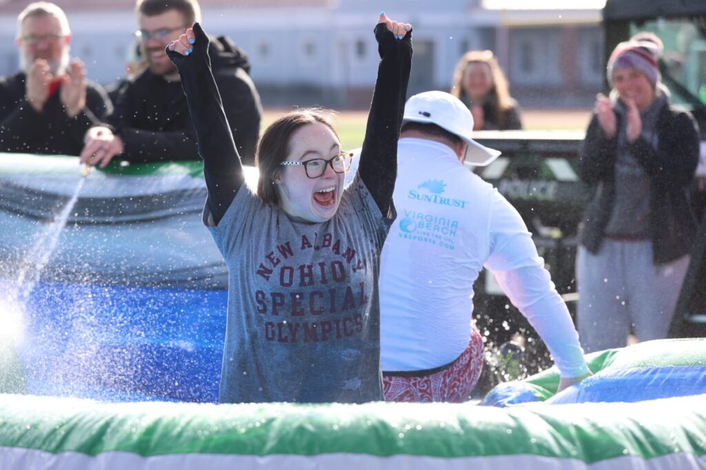 Woman in glasses wearing a wet New Albany Ohio Special Olympics shirt raises her arms in an inflatable pool as people clap.