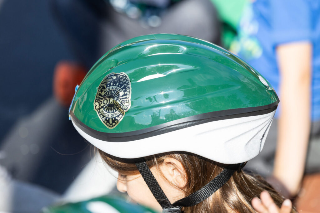 Child wearing green-and-white bicycle helmet with a "Police Junior Officer" badge sticker, black chin strap visible