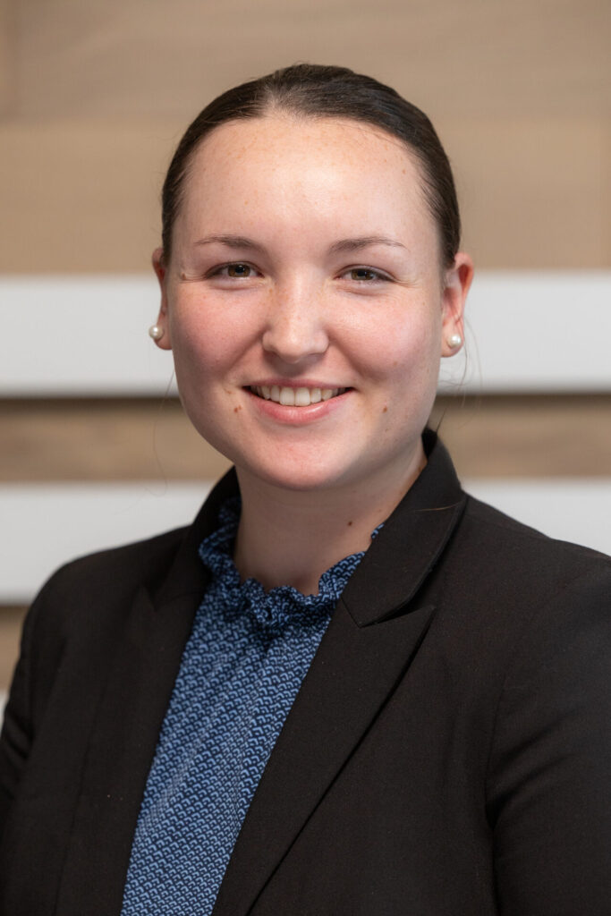 Young woman with hair pulled back, wearing pearl earrings, a blue patterned blouse and black blazer, smiling at the camera.