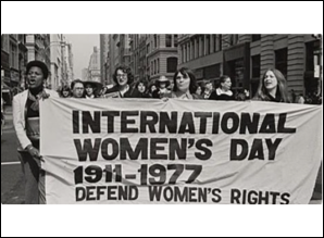 Group of women marching in a city street holding a large banner reading "INTERNATIONAL WOMEN'S DAY 1911-1977 DEFEND WOMEN'S RIGHTS."