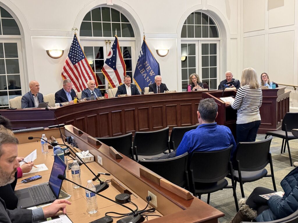 Woman addressing a seated municipal board at a curved wooden dais with American and state flags behind the panelists.
