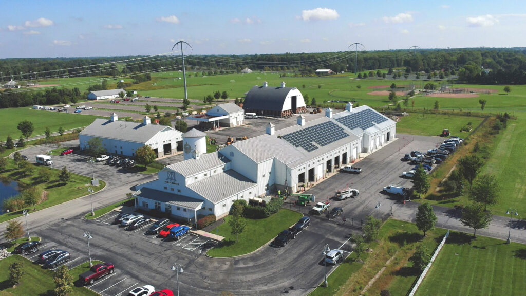 Aerial view of a white service complex with solar panels on the roof, adjacent parking lots and green sports fields beyond