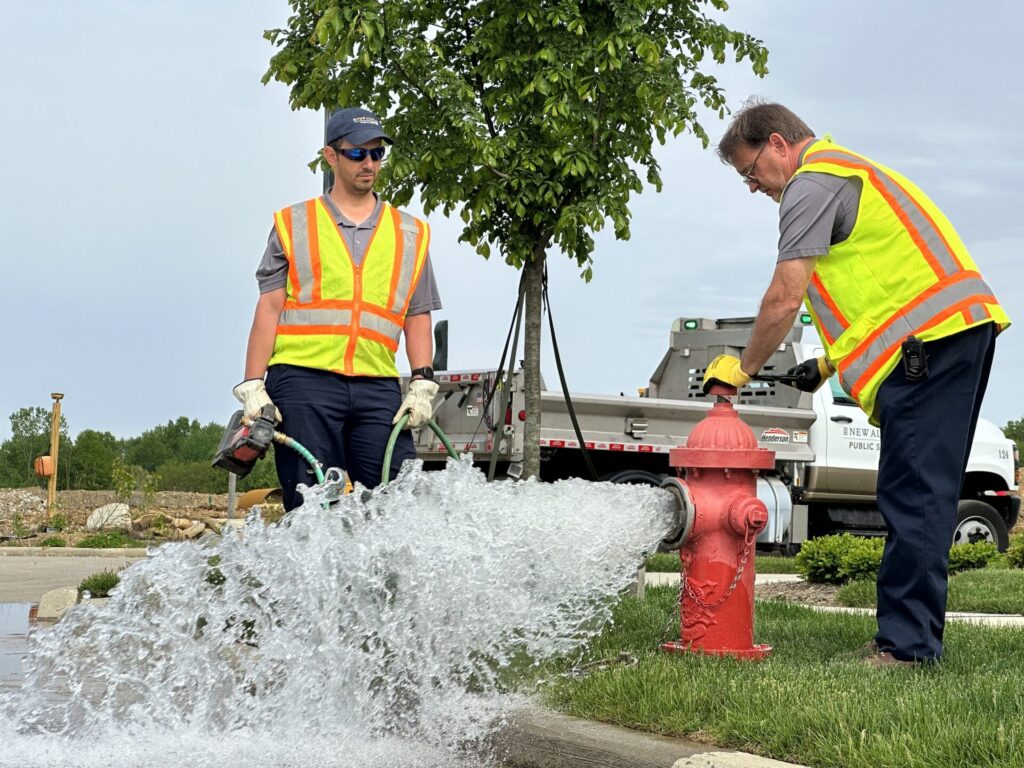Two workers in safety vests supervising a red fire hydrant gushing water onto a street during outdoor maintenance