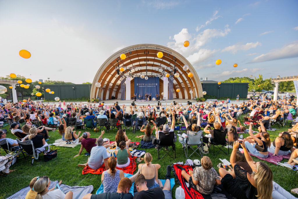 Outdoor concert at the Hinson Amphitheater with a large seated crowd tossing yellow balloons into the sky.
