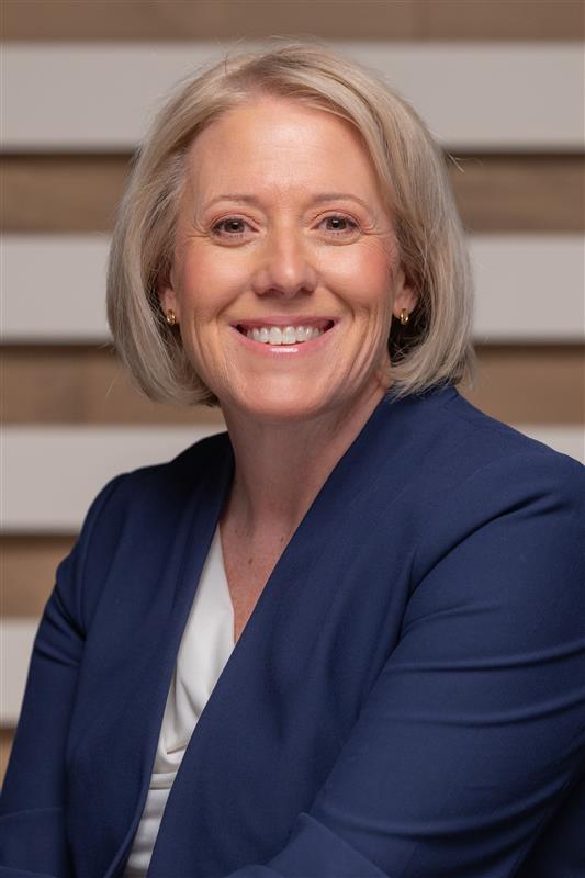 Smiling middle-aged woman with short blonde hair wearing a navy blazer and white blouse, seated against a striped wood background