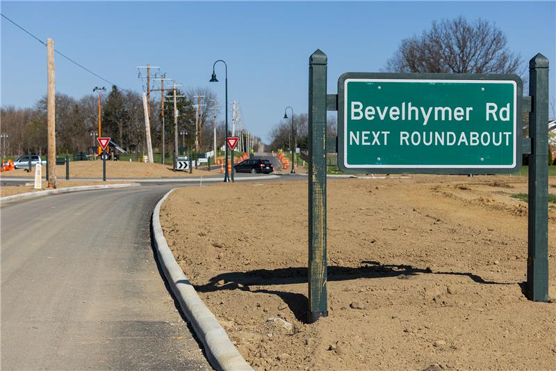 Green road sign reading "Bevelhymer Rd NEXT ROUNDABOUT" beside a newly paved road and bare dirt median with roundabout ahead