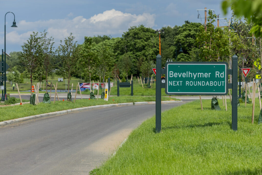 Green road sign reading Bevelhymer Rd NEXT ROUNDABOUT beside a paved road with young trees and grassy median indicating the Bevelhymer Road and Walnut Street roundabout