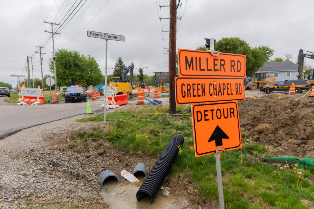 Orange detour signs reading Miller Rd and Green Chapel Rd at a busy roadside construction site with equipment and police cars