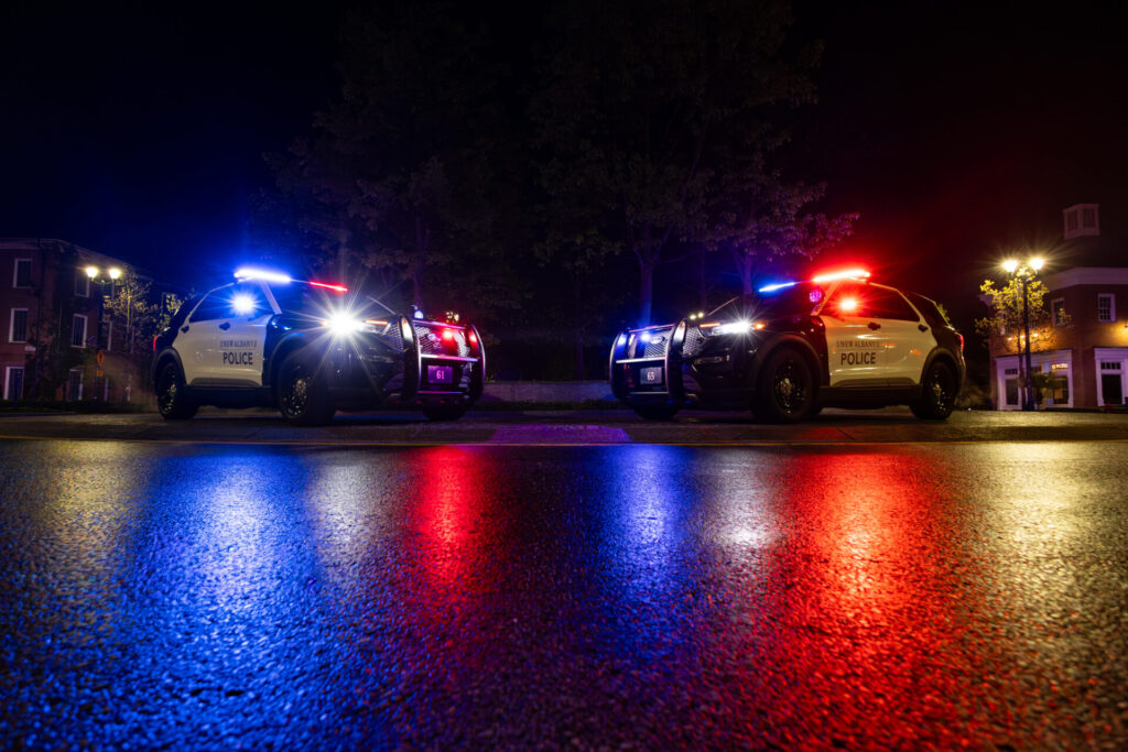 Two parked New Albany police SUVs facing each other at night with red and blue lights reflecting on wet pavement.