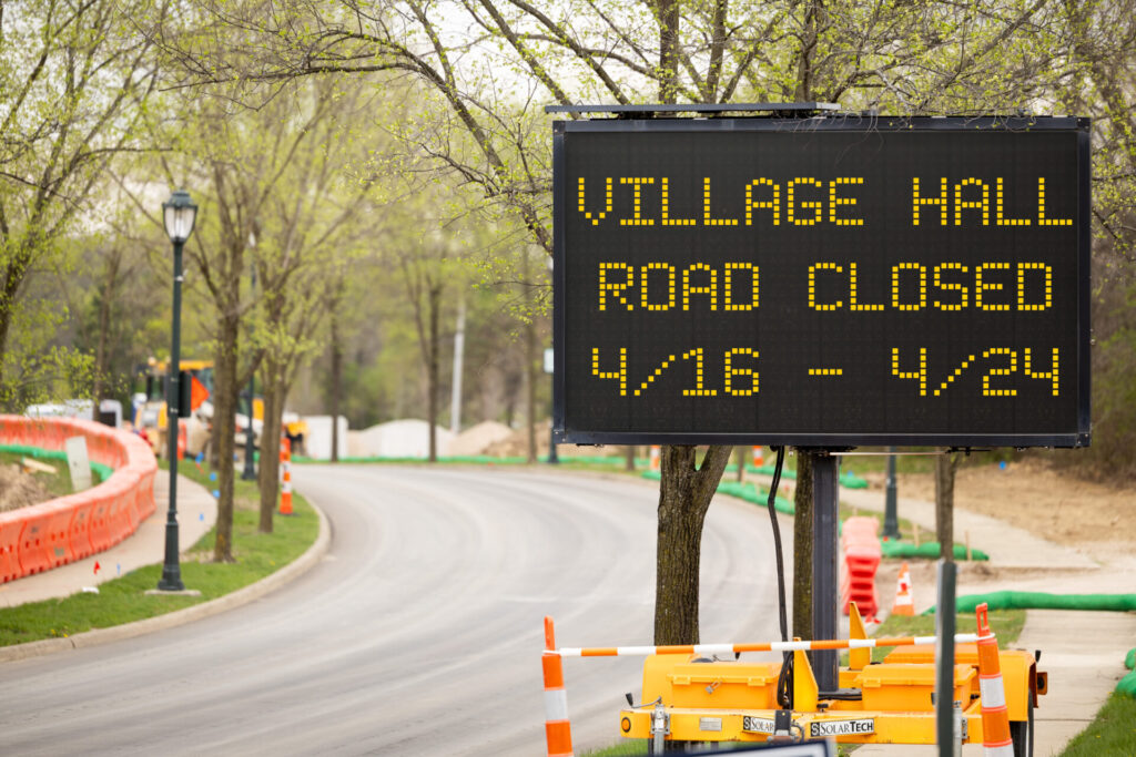 Electronic road sign beside a curved tree-lined street reading "VILLAGE HALL ROAD CLOSED 4/16 - 4/24" with construction barriers nearby