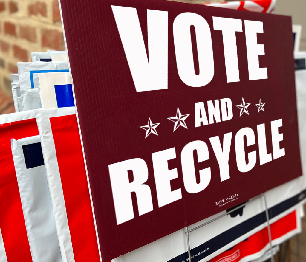 Stacked lawn signs with a large maroon sign reading "VOTE AND RECYCLE" in white letters and star graphics.