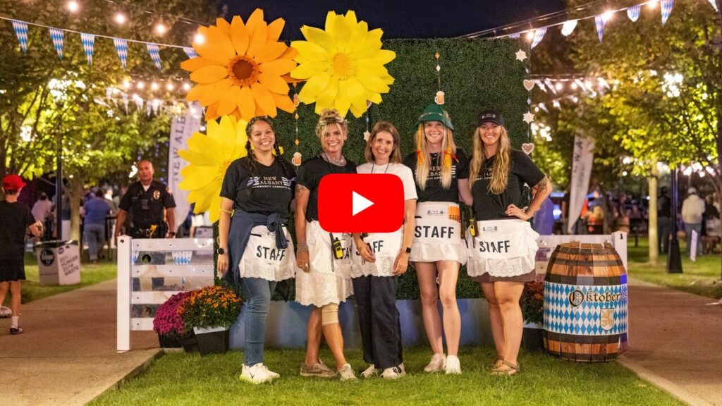 Five smiling festival staff wearing white aprons pose under large decorative sunflowers and string lights at night.