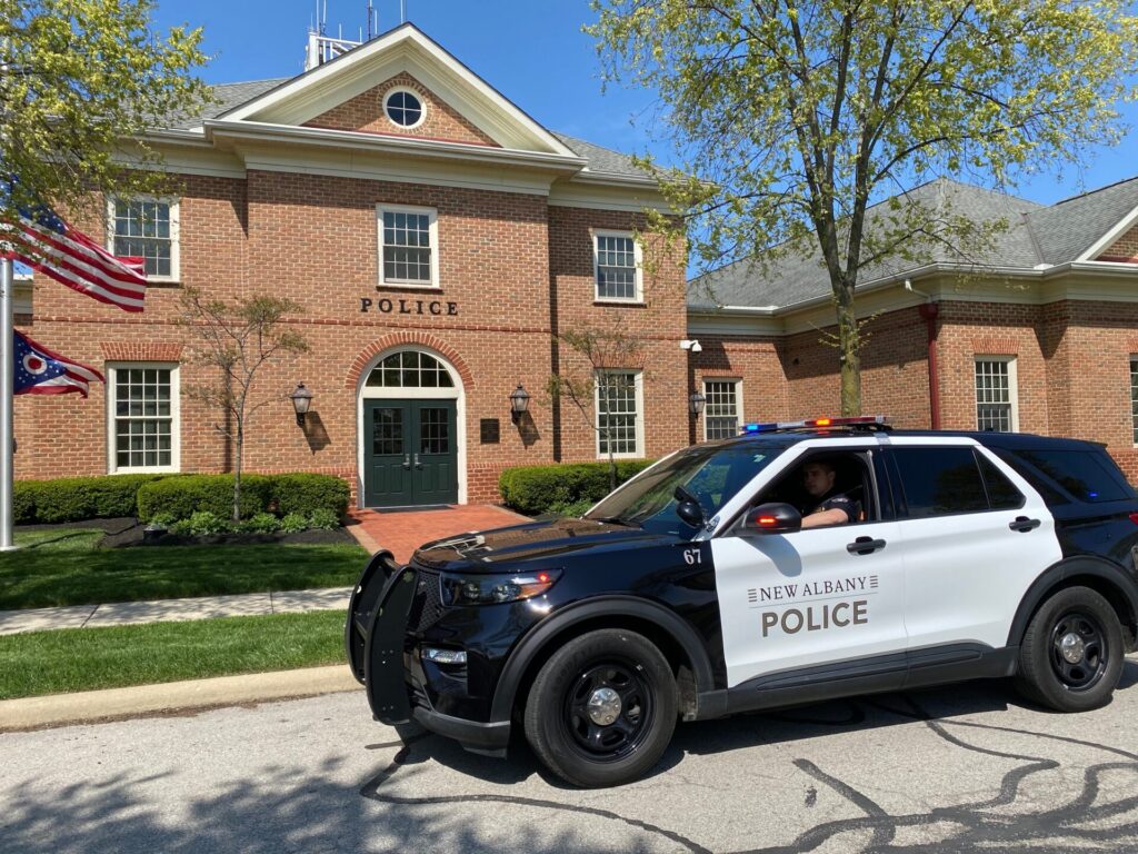 Black and white New Albany police SUV with lights on parked in front of a brick police station building with flags and trees.