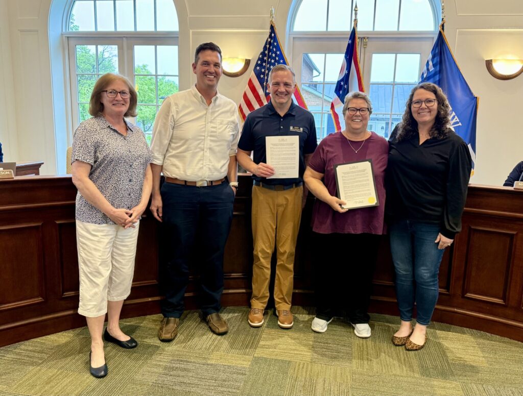 Five smiling adults standing in a row inside a government chamber, two holding framed certificates, U.S. and state flags behind them.