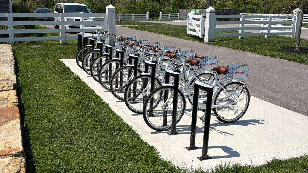 Row of white rental bicycles with brown seats docked at a bike rack on a sidewalk beside a grassy area and white fence