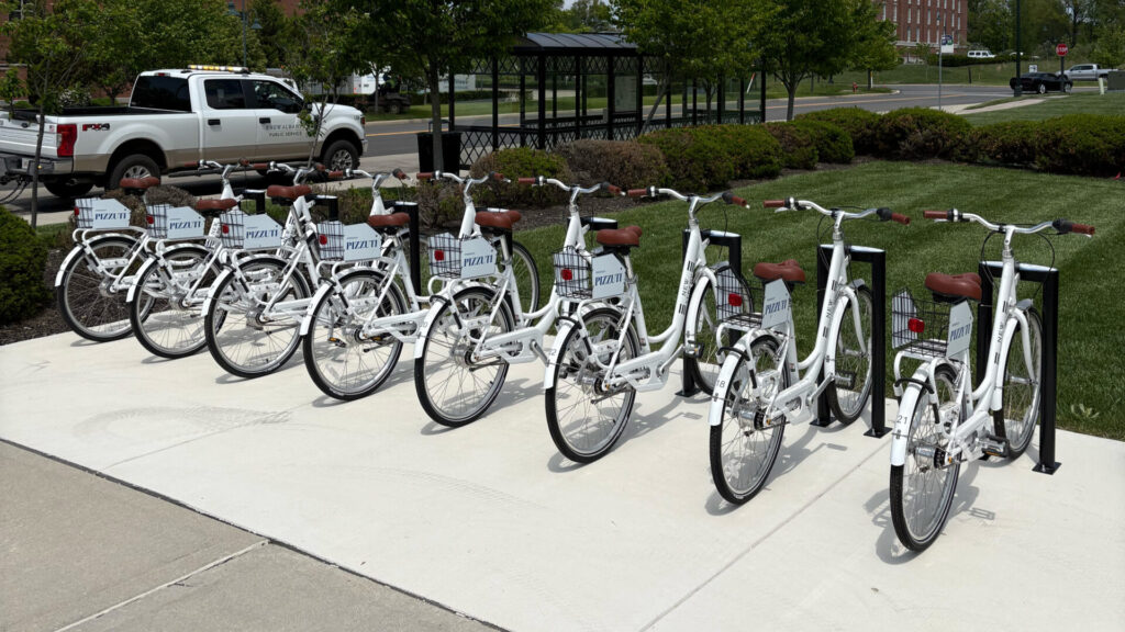 Row of white rental bicycles with brown seats and front baskets labeled "PIZZUTI" parked at a sidewalk docking station