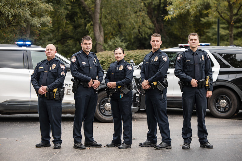 Five uniformed police officers standing in a line in front of two police SUVs with lights on, outdoors with trees behind them.