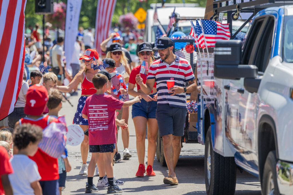 Crowd of people in red, white and blue passing out small American flags and treats from a decorated truck during a patriotic parade.