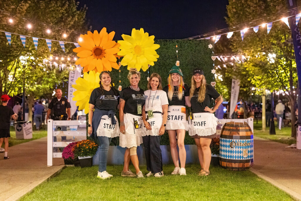 Five smiling New Albany event staff wearing aprons and festival shirts pose in front of giant paper sunflowers and string lights at night