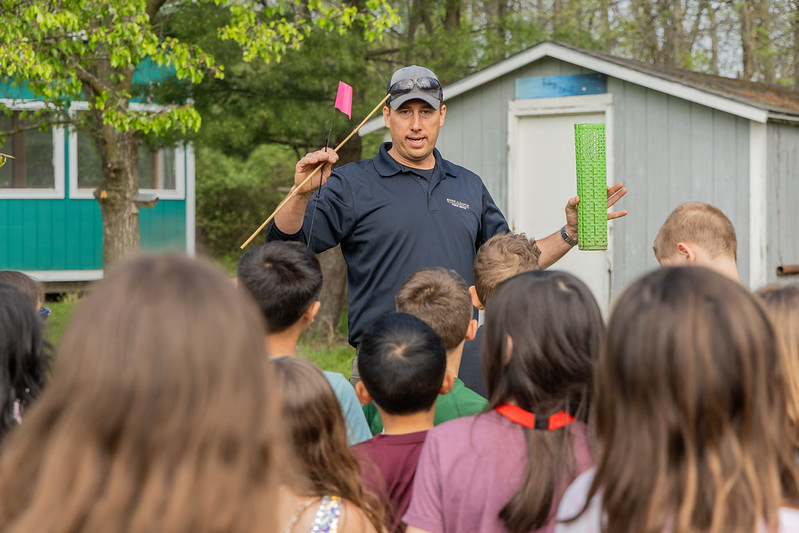 Man in cap demonstrating a green tube and flag to a group of children outdoors near small sheds.