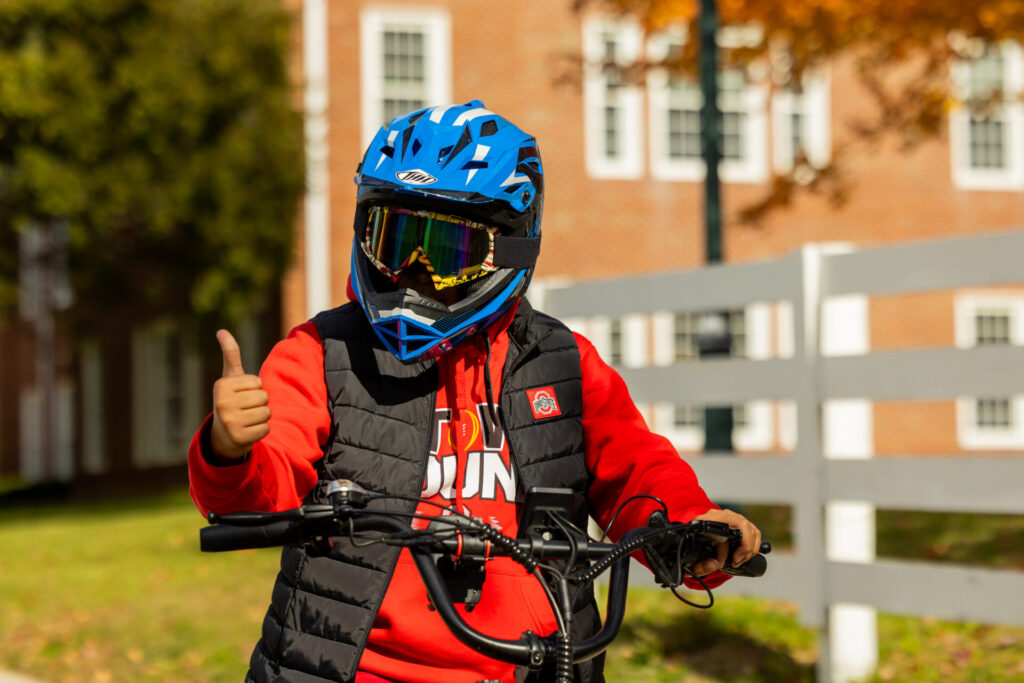 Person wearing a blue full-face helmet and mirrored goggles on a bike, giving a thumbs-up while wearing a red hoodie and black vest