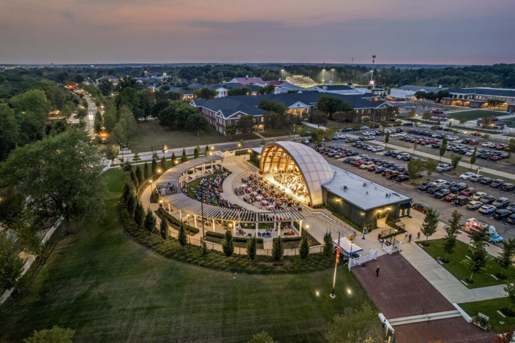Hinson Amphitheater at dusk with a lit stage and seated audience, surrounded by parking lots, lawns, and nearby buildings