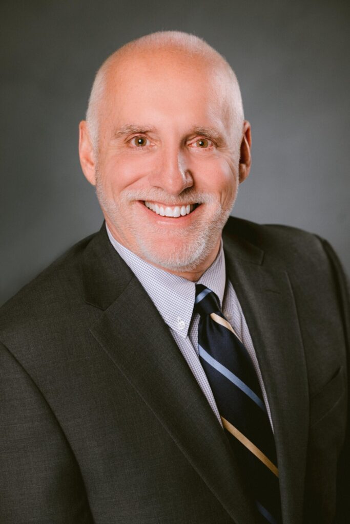 Smiling bald middle-aged man with trimmed white beard wearing a dark suit, checkered shirt and striped tie against a gray background
