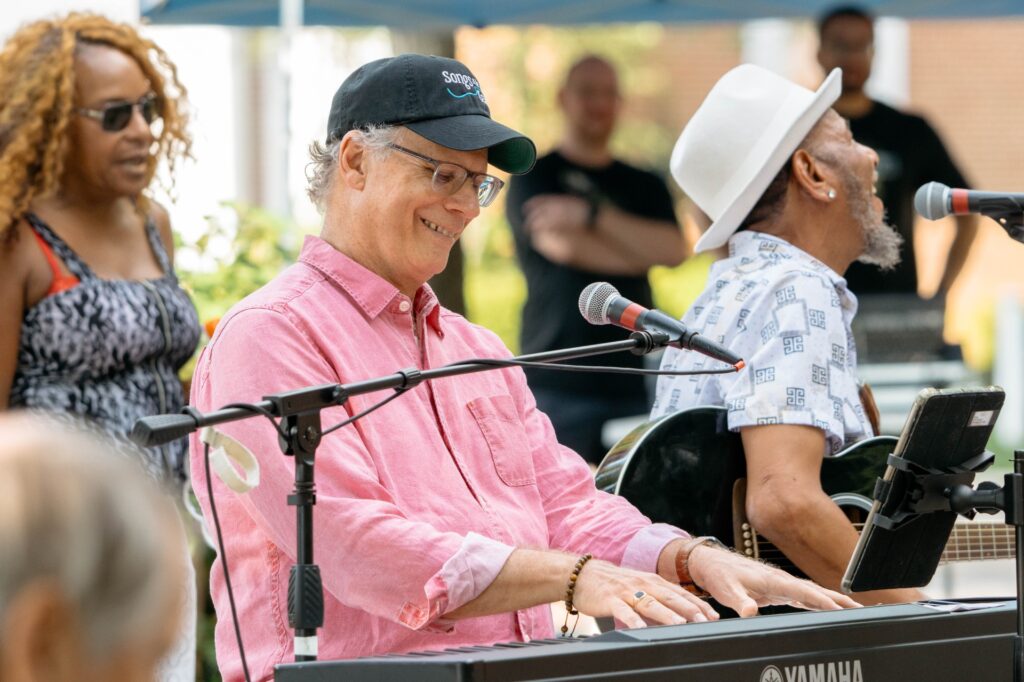 Eric Gnezda in a pink shirt and black cap smiling while playing a keyboard beside a guitarist in a white hat and a backing vocalist.