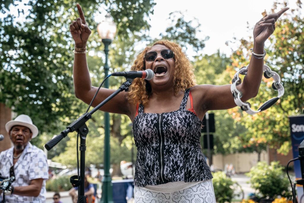 Black woman singer with sunglasses and tambourine raised, mouth open into a microphone during an outdoor performance.