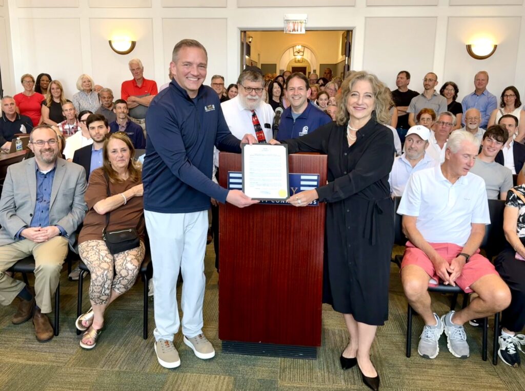 Two smiling adults at a podium jointly holding a framed certificate while a crowded audience watches in a room.