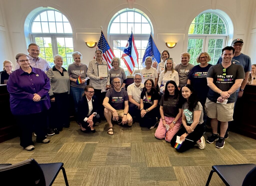 Diverse group of people posing indoors with flags and rainbow pride items, some wearing "Love Thy Neighbor" shirts.
