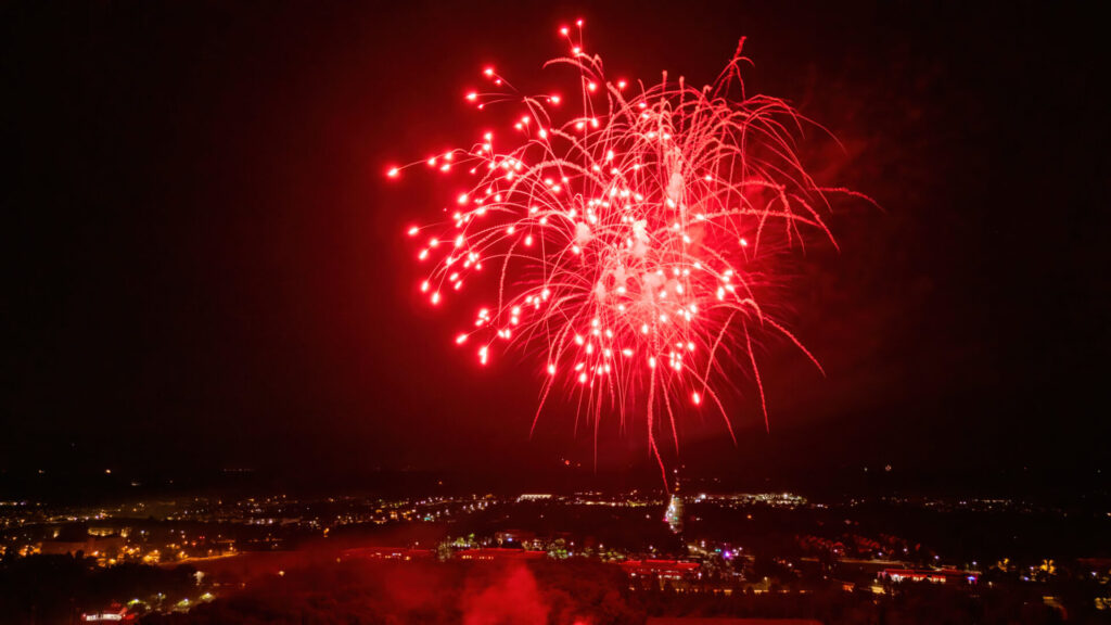 Bright red fireworks burst over a lit town at night, streams and glowing sparks fanning outward above the skyline.