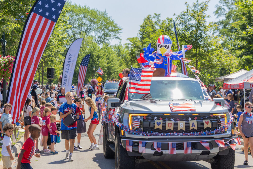 Parade scene with a decorated pickup covered in American flags and a large inflatable eagle, surrounded by people.